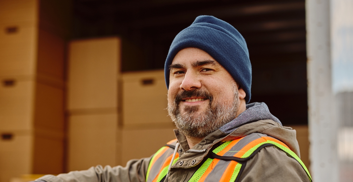 Dock worker wearing a blue beanie and high-visibility safety vest, standing in front of stacked cardboard boxes at a loading dock