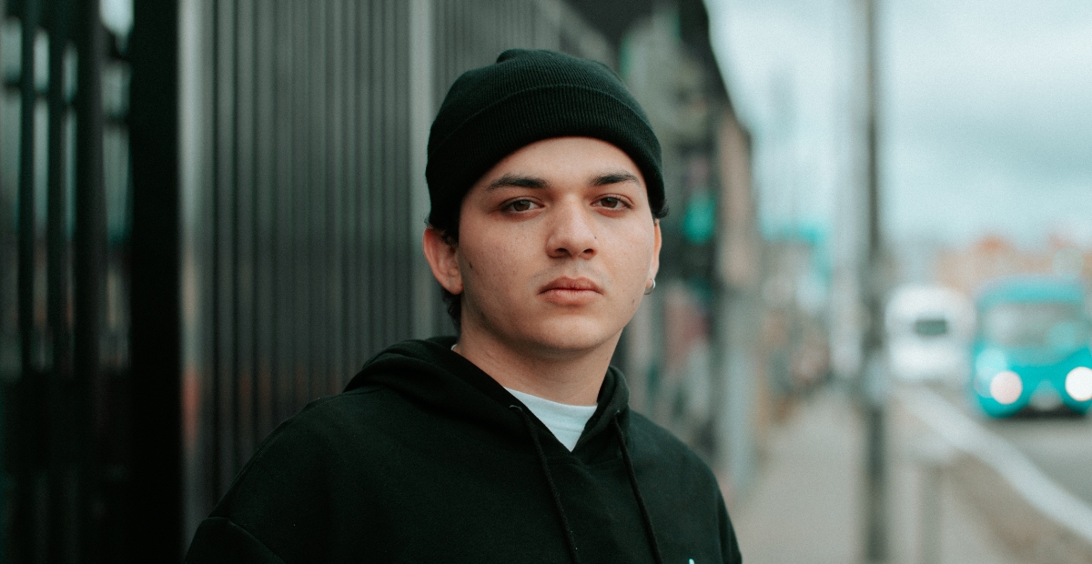 A young person wearing a black beanie and a black hoodie stands outdoors near a blurred street background.