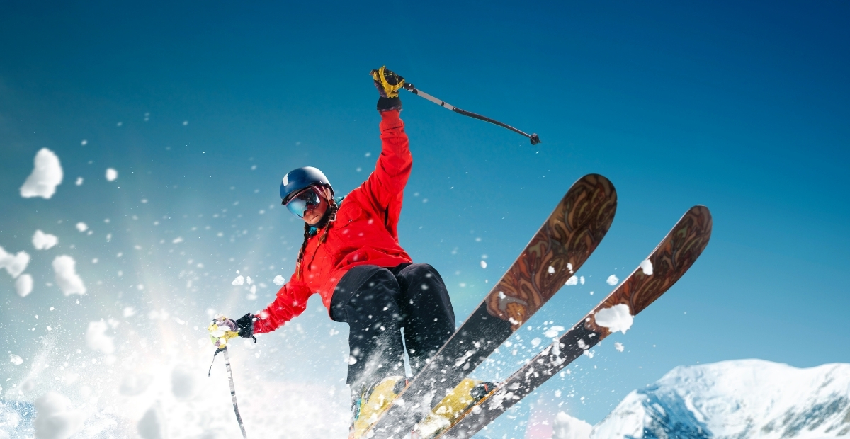 Ein Skifahrer in roter Jacke und Helm springt mit nach oben geneigten Skiern von einem verschneiten Hang ab, während um ihn herum Schnee aufwirbelt und der Himmel strahlend blau ist.