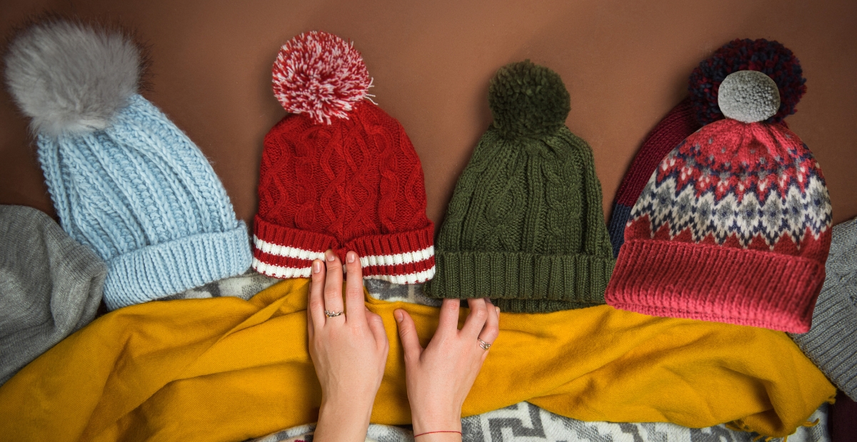 A row of colorful knit beanies with pom-poms laid out on a surface, including light blue, red with white stripe, olive green, and a patterned red-blue-pink pom-pom beanie. A mustard yellow blanket is partially visible in the foreground.