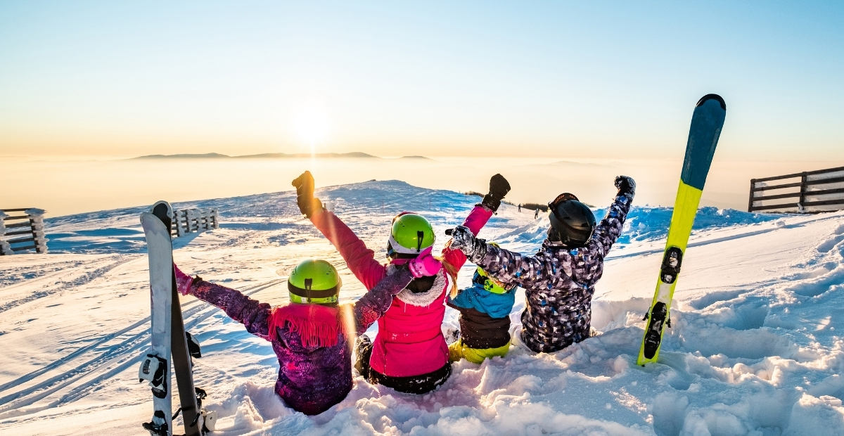A group of four children sit in the snow at a sunny ski slope, wearing colorful winter jackets and helmets, with skis standing upright nearby.