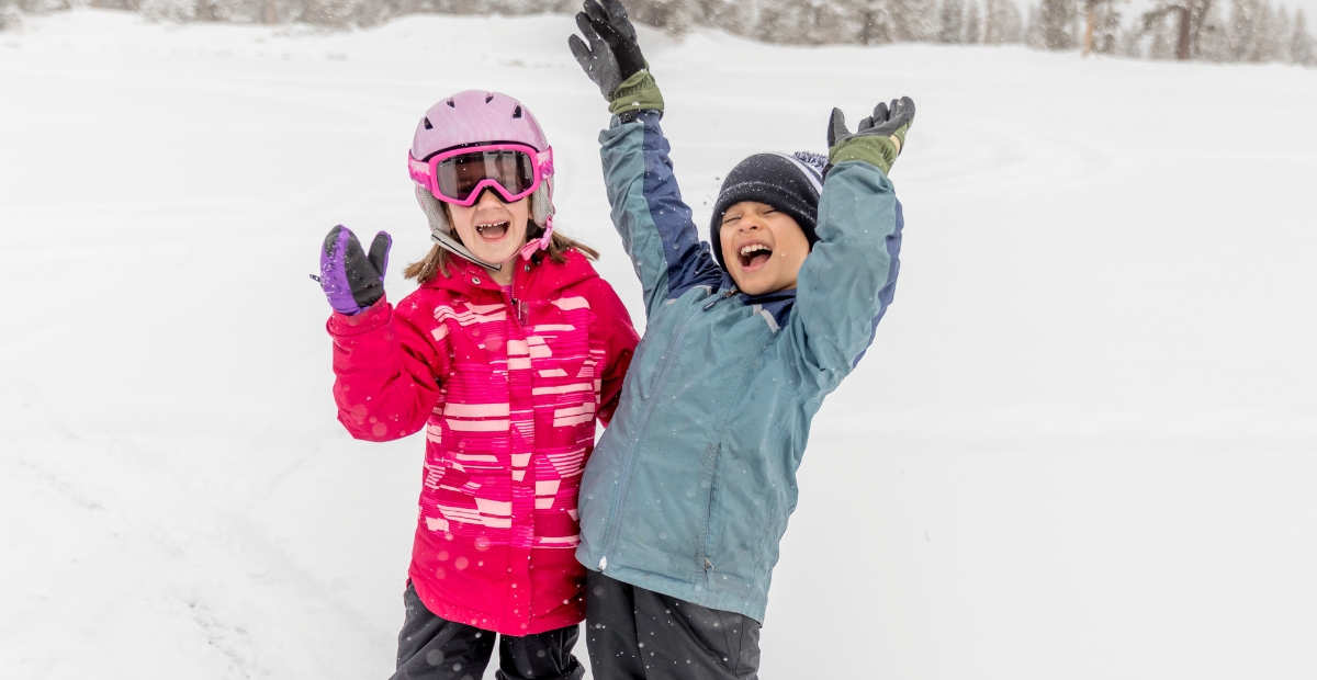 Two children in colorful winter ski outfits stand in the snow, smiling and raising their gloved hands in celebration. The girl wears a pink helmet and jacket, the boy a blue jacket and dark knit hat.