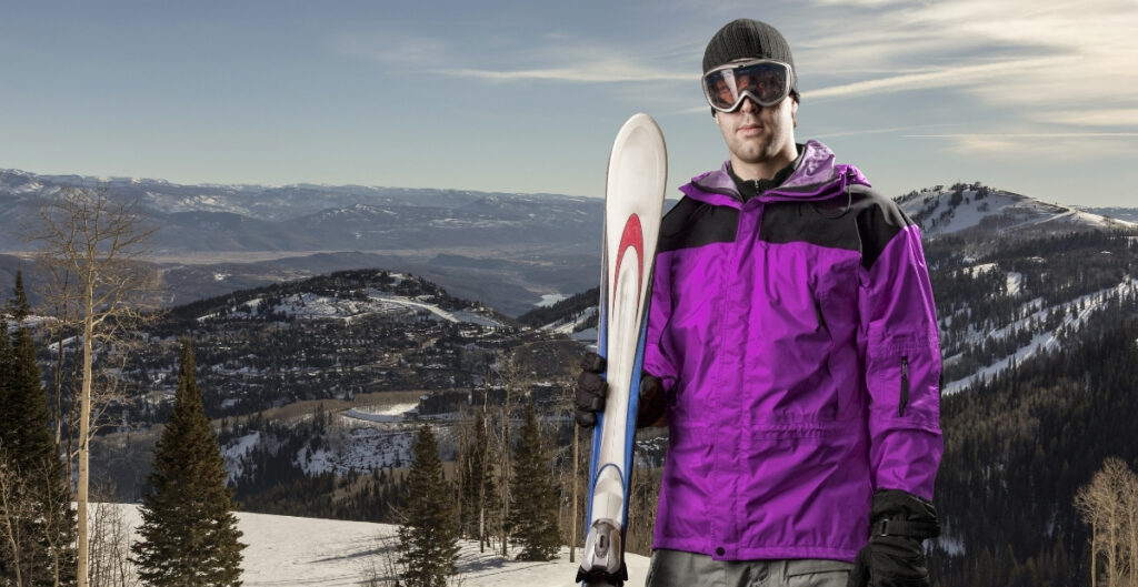 A man dressed in a bright purple ski jacket and dark pants stands on a snowy mountainside, holding a snowboard and wearing goggles and a beanie with a wintry alpine landscape in the background.
