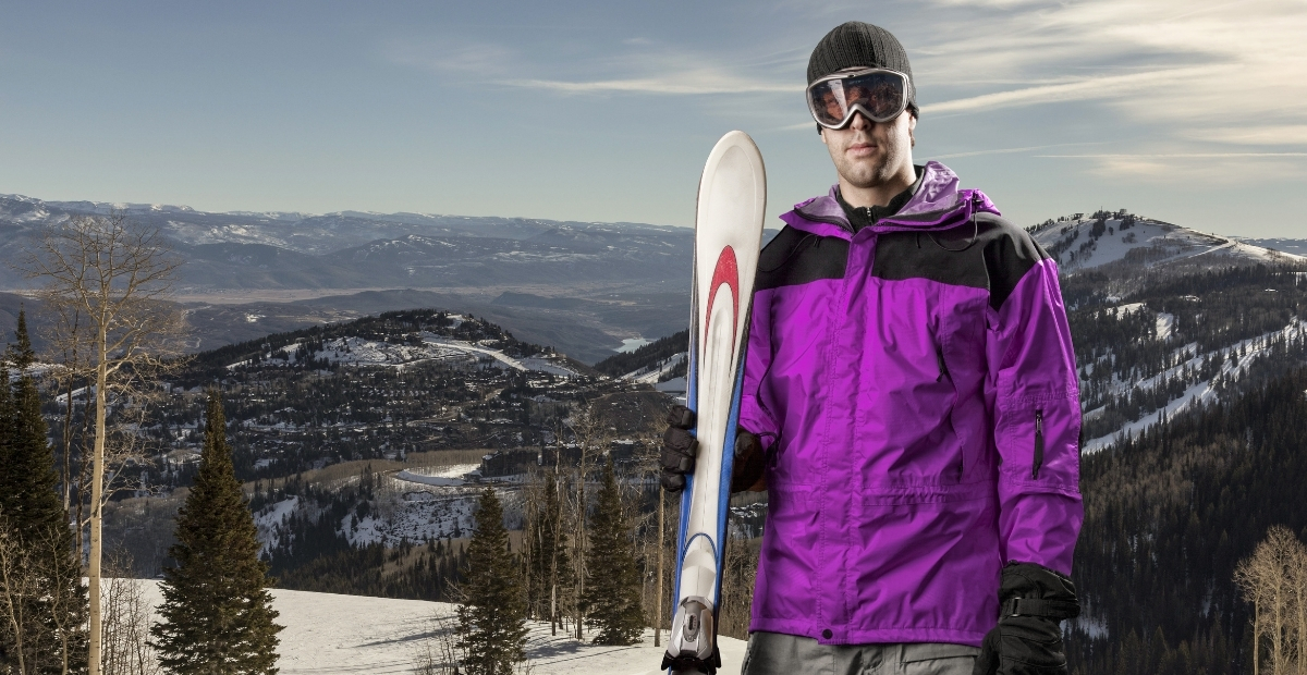 A man dressed in a bright purple ski jacket and dark pants stands on a snowy mountainside, holding a snowboard and wearing goggles and a beanie with a wintry alpine landscape in the background.