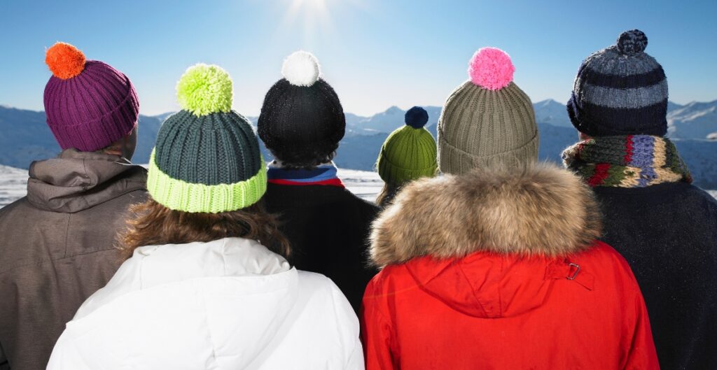 Group of people from the back wearing colorful retro-style knit beanies with large pom-poms, snow-capped mountains in the background.
