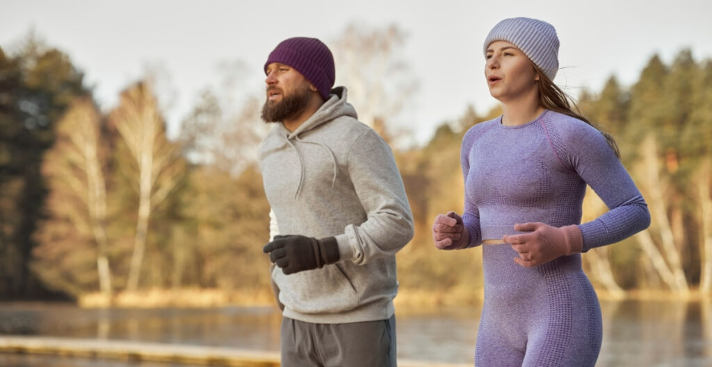 A man and a woman running outdoors by a lake, both wearing knit beanies and activewear in cool weather.