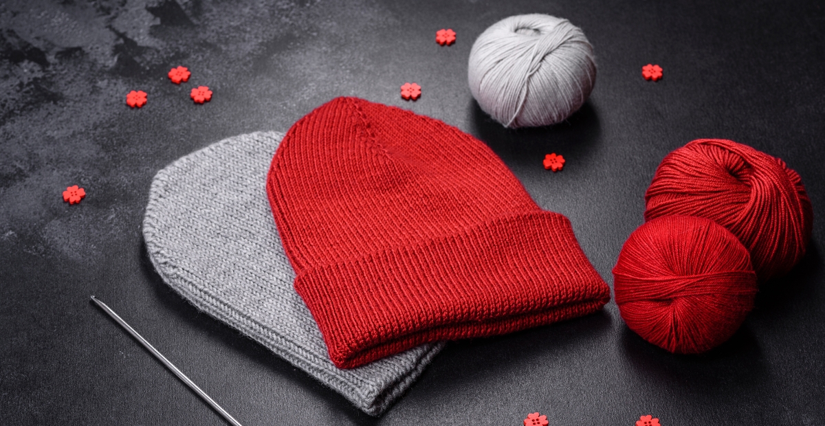A red seamless knit beanie rests on top of a light gray seamless knit beanie, with balls of red and gray yarn and a knitting needle scattered around on a dark surface.