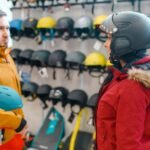 A man and a woman stand in a ski shop. The man on the left wears a yellow and orange jacket and holds a blue ski helmet. The woman on the right wears a red jacket with a fur-lined hood and a dark ski helmet. Behind them is a wall display of helmets in various colors and designs, with skis and other gear visible on lower shelves.