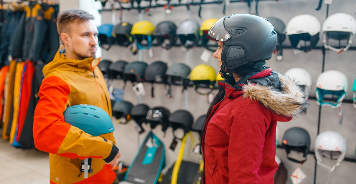 A man and a woman stand in a ski shop. The man on the left wears a yellow and orange jacket and holds a blue ski helmet. The woman on the right wears a red jacket with a fur-lined hood and a dark ski helmet. Behind them is a wall display of helmets in various colors and designs, with skis and other gear visible on lower shelves.