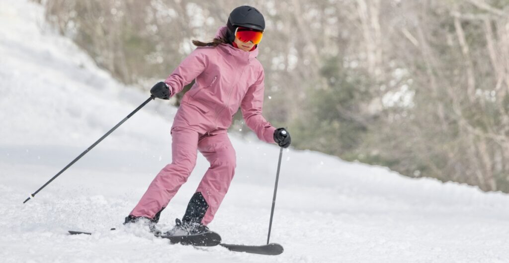 A person skiing down a snowy slope wearing a pink ski suit, black helmet, and red goggles, holding ski poles with visible powder spray.