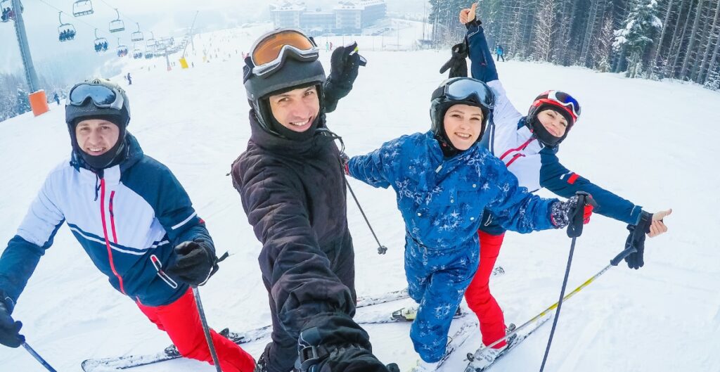 A group of four friends in ski gear posing cheerfully on a snowy slope, wearing helmets, goggles, and colorful ski outfits with ski poles. A ski lift and snowy trees are visible in the background.