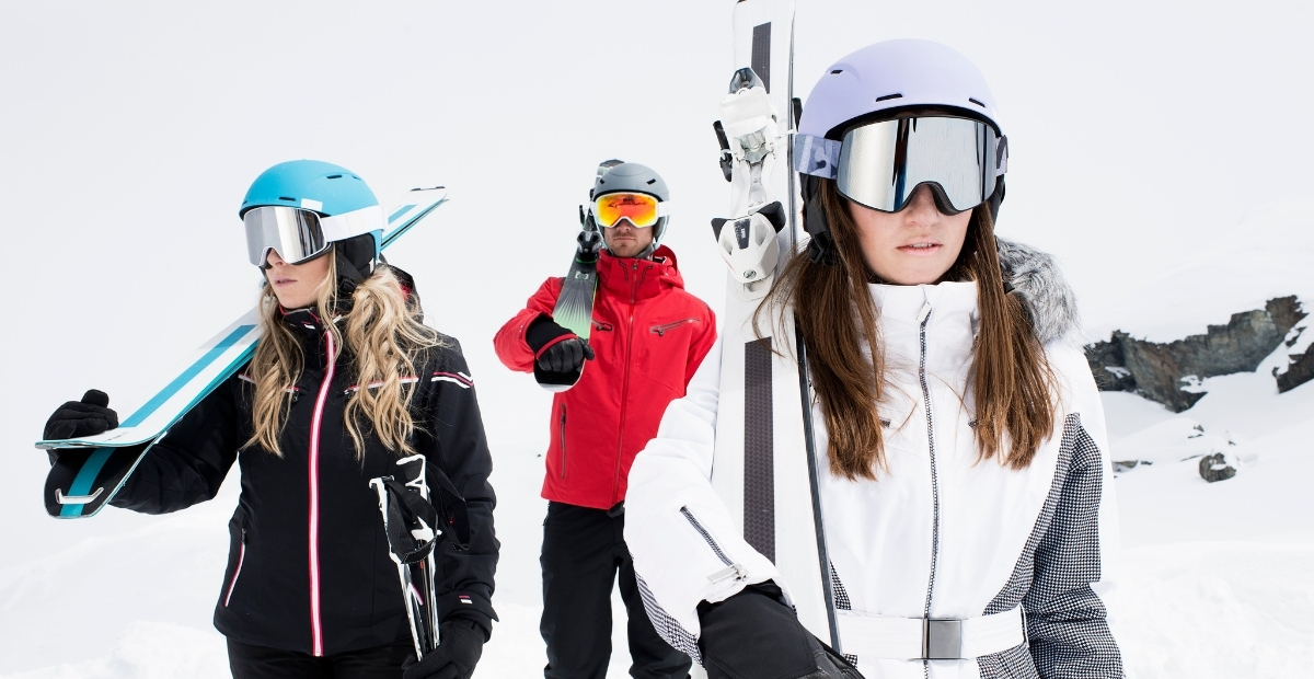Three skiers in winter gear standing in a snowy landscape: two women in the foreground with helmets and ski goggles, one carrying skis, and a man in a red jacket in the background holding ski poles.