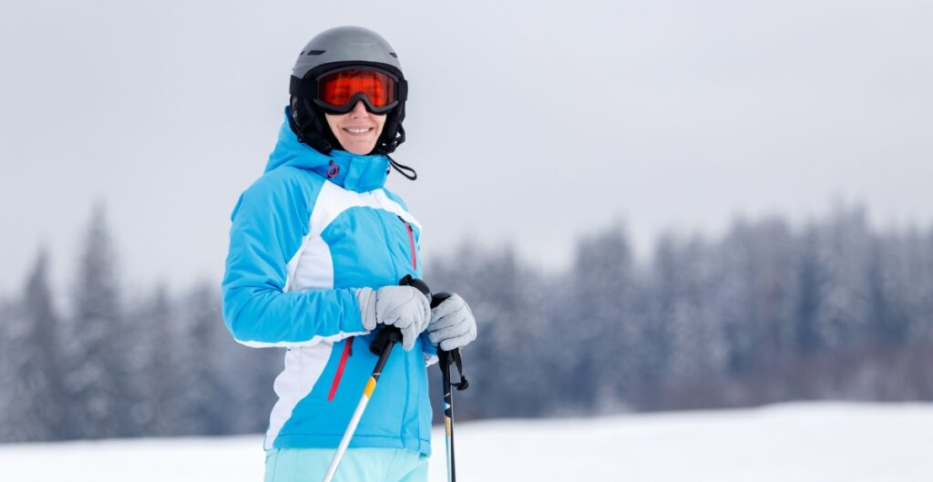 A woman in ski gear stands in a snowy landscape, smiling at the camera. She wears a grey helmet, orange ski goggles, and a blue and white ski jacket, with light gloves and holding ski poles. Snow-covered ground and a blurred treeline under a grey sky are in the background.