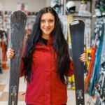 A woman with long dark hair wearing a red jacket stands in a ski shop, smiling while holding a pair of dark skis. Ski racks and winter clothing are visible in the background.
