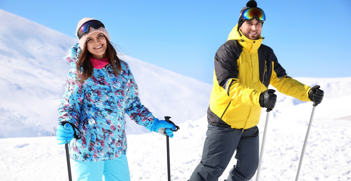 A man and a woman stand on a snowy mountain slope, both dressed in colorful ski gear and holding ski poles. The woman wears a blue and pink patterned jacket with light blue pants, a beige knit hat, and ski goggles on her forehead, smiling at the camera. The man wears a yellow and black jacket with gray pants and black gloves, wearing ski goggles and a black beanie. Bright blue sky and snowy mountains in the background.