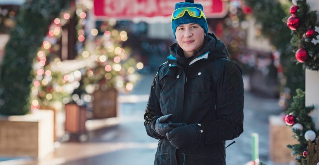 A person dressed in winter ski wear stands outdoors on a street decorated with Christmas lights and trees. They wear a black jacket, blue knit hat, and ski goggles resting on their forehead.