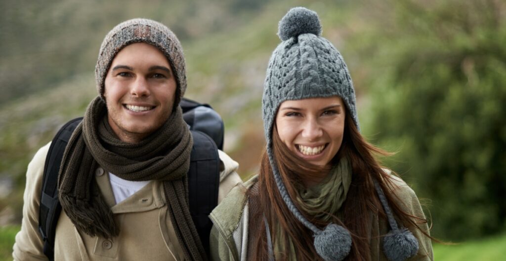 "Two smiling people outdoors wearing knit beanies and scarves, dressed for cool weather."