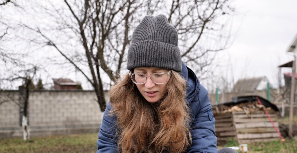 A young woman outdoors wearing a gray knit beanie, round clear-framed glasses, and a blue puffer jacket. She has long wavy hair and is looking downward, with a bare tree and a residential area in the background.