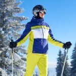 Un esquiador se encuentra de pie en una pista nevada con una chaqueta azul y amarilla, pantalones amarillos brillantes, casco y gafas protectoras, sosteniendo unos bastones de esquí, con árboles cubiertos de nieve y un cielo azul claro al fondo.