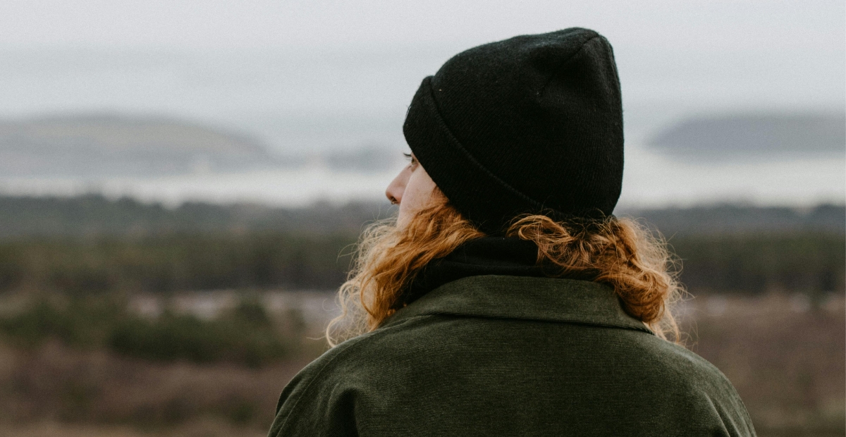 A person with wavy hair wearing a black knit beanie and a dark green jacket, photographed from behind in an outdoor setting with a blurred landscape in the background