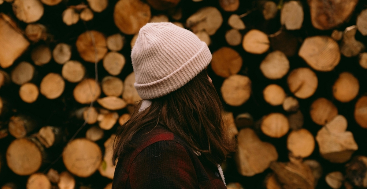 A person with long dark hair wearing a beige knit beanie and a dark plaid jacket, standing in front of a large stack of cut log rounds.