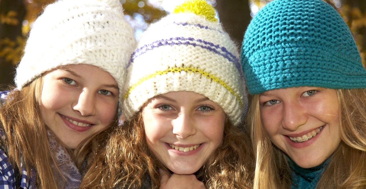 Three smiling girls wearing chunky waffle-knit beanies: white on the left, white with colorful stripes in the middle, and turquoise on the right, outdoors with autumn light.