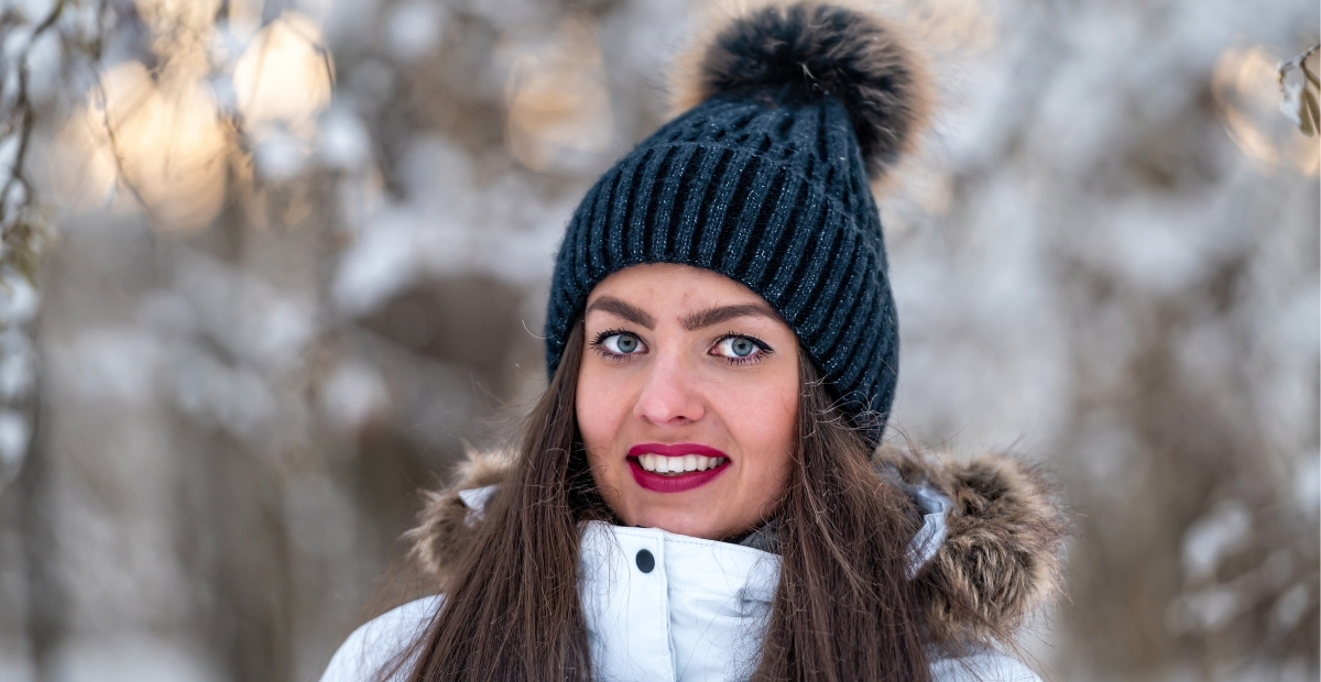 A woman wearing a dark blue knit beanie with a fluffy pom-pom, smiling outdoors in a snowy, blurred forest background.