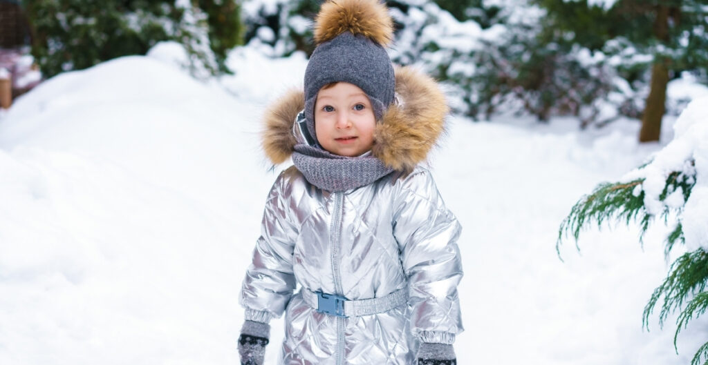 A young child standing in the snow wearing a metallic silver puffer ski suit with a fur-lined hood, gray knit hat with a pom-pom, scarf, and gloves.