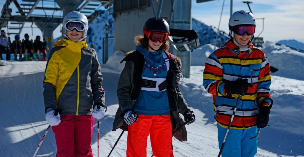 Three teenagers stand on a snowy ski slope wearing colorful ski outfits with helmets and goggles, each holding ski poles. A ski lift and snowy mountains are in the background.
