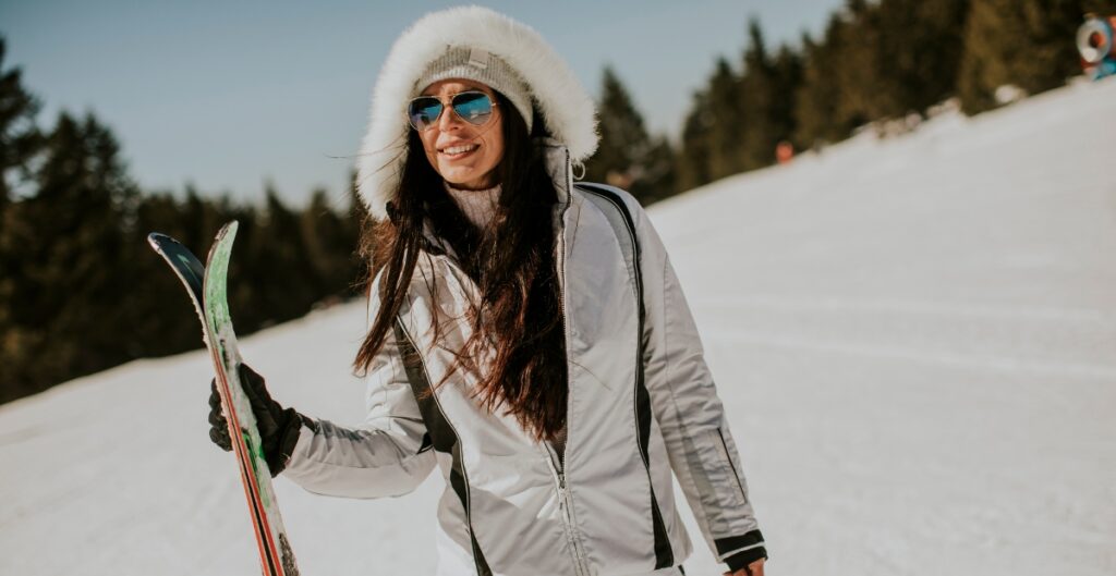 A woman on a snowy slope wearing a white winter ski jacket with a fur-lined hood and reflective sunglasses, holding skis and smiling. Evergreen trees and a clear blue sky are in the background.