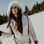 Una mujer en una pista nevada con una chaqueta de esquí blanca con capucha forrada de piel y gafas de sol reflectantes, sosteniendo unos esquís y sonriendo. Al fondo se ven árboles de hoja perenne y un cielo azul claro.