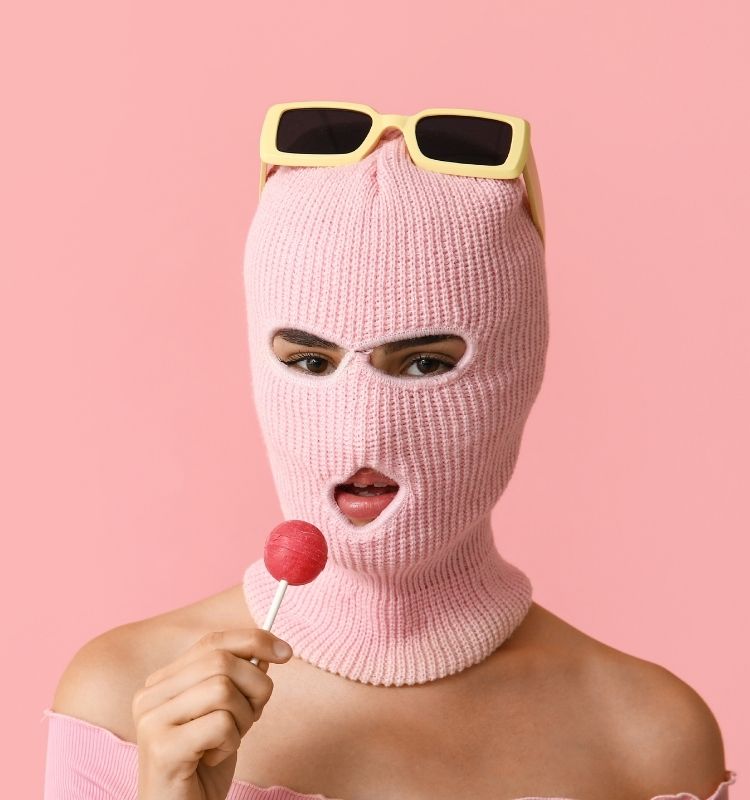 Woman wearing a pink knitted balaclava with yellow sunglasses, holding a red lollipop, against a pastel pink background.