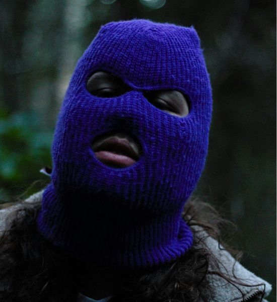 A person wearing a vibrant pink knitted balaclava with holes for eyes and mouth, adorned with a safety pin and a shiny silver chain, paired with a black blazer.