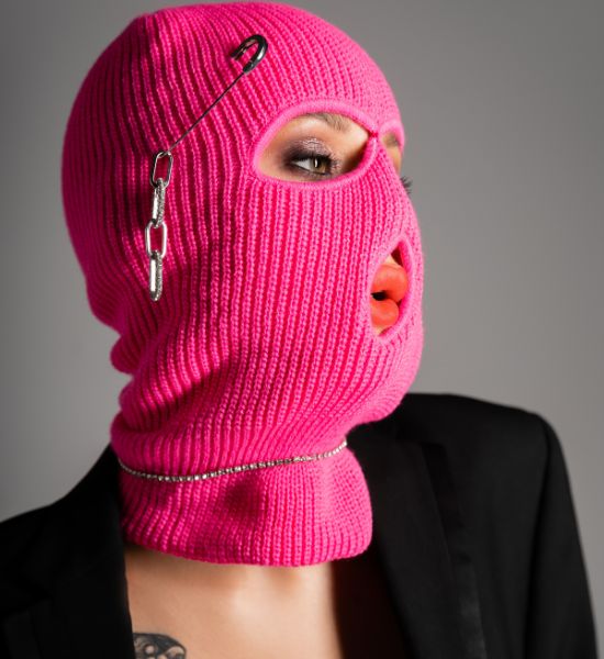 A close-up of a person wearing a bright pink knitted balaclava with safety pins and a silver chain, contrasted against a black blazer.