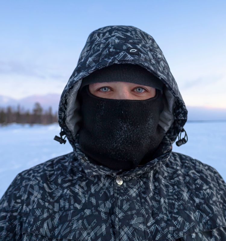 Person wearing a black thermal balaclava and winter jacket in extreme cold snowy landscape.