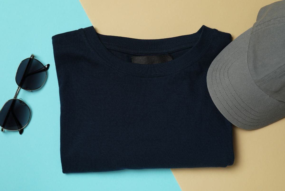 A flat lay of a navy blue T-shirt, a gray cap, and black sunglasses arranged on a background split into beige and light blue sections.