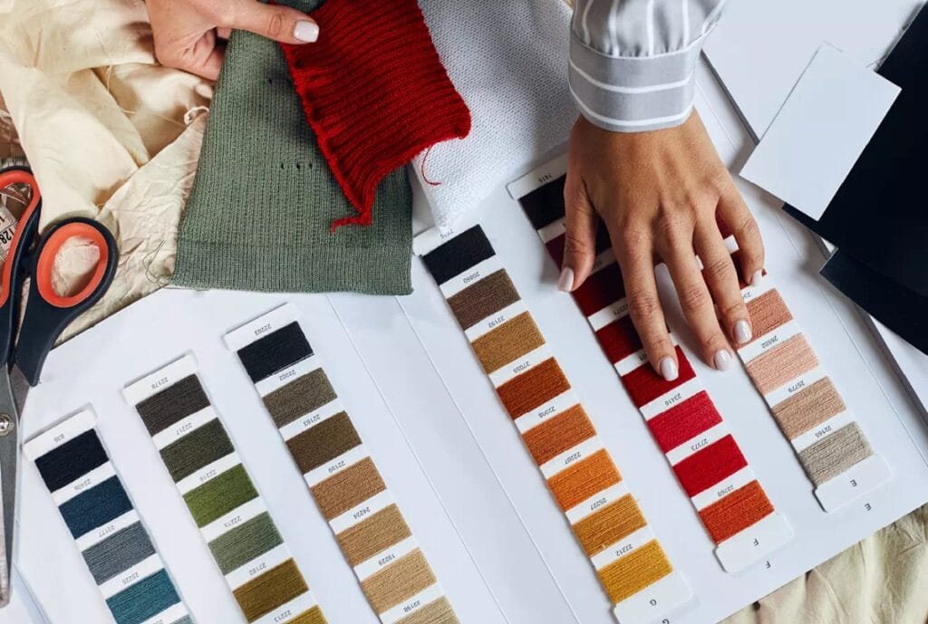 A person’s hand selecting a red fabric swatch amidst an array of color sample cards, with sewing scissors, fabric pieces, and color strips laid out on a table.