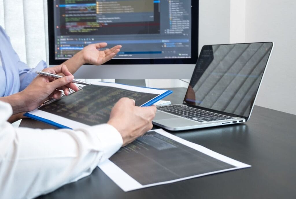 Two professionals working together at a desk, reviewing digital code on a tablet, with a laptop and large monitor displaying programming environments.