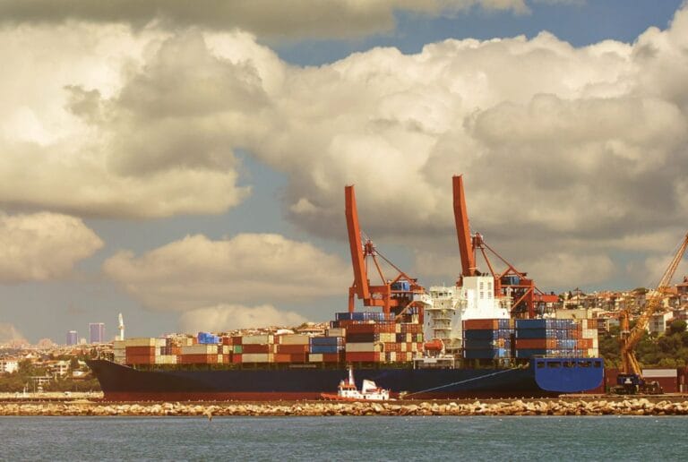 Container ship being loaded by two red cranes at a port under a cloudy sky