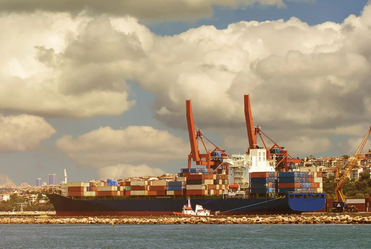 A large cargo ship carrying numerous stacked shipping containers, docked at a port with cranes in the background and a city skyline under a partly cloudy sky.