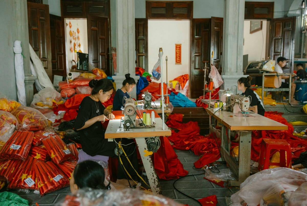 Workers at sewing machines in a busy workshop surrounded by red fabrics and materials, illustrating in-house manufacturing technology.