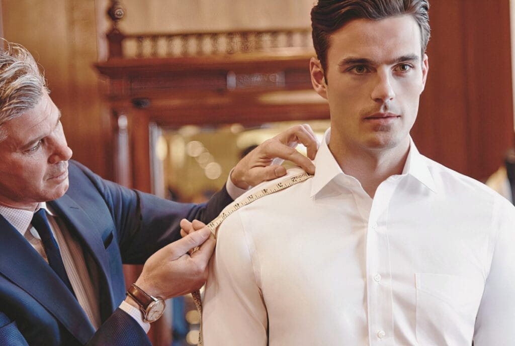 A tailor or fit specialist measuring the shoulder of a young male model wearing a white dress shirt in a fitting room or tailoring shop.
