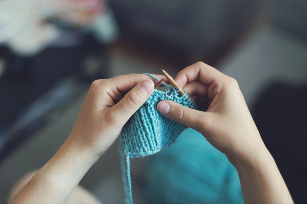 A person knitting with turquoise yarn, holding knitting needles and working on a small piece of fabric.