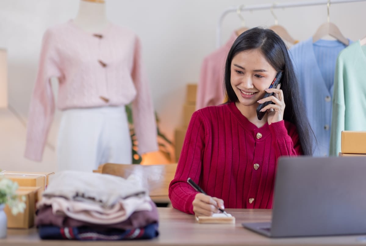 A woman working at a clothing store or showroom, talking on the phone, with folded clothes and clothing racks in the background.