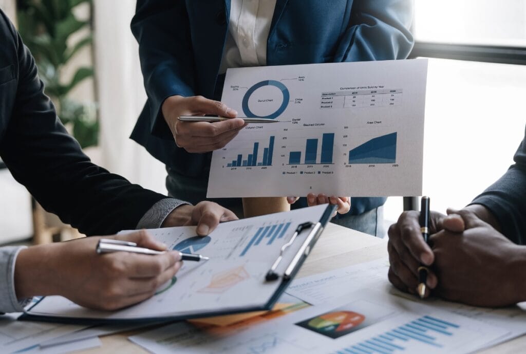 Three business professionals engaged in a discussion, with one person holding a chart displaying various graphs and data, while the others take notes and review documents on the table.