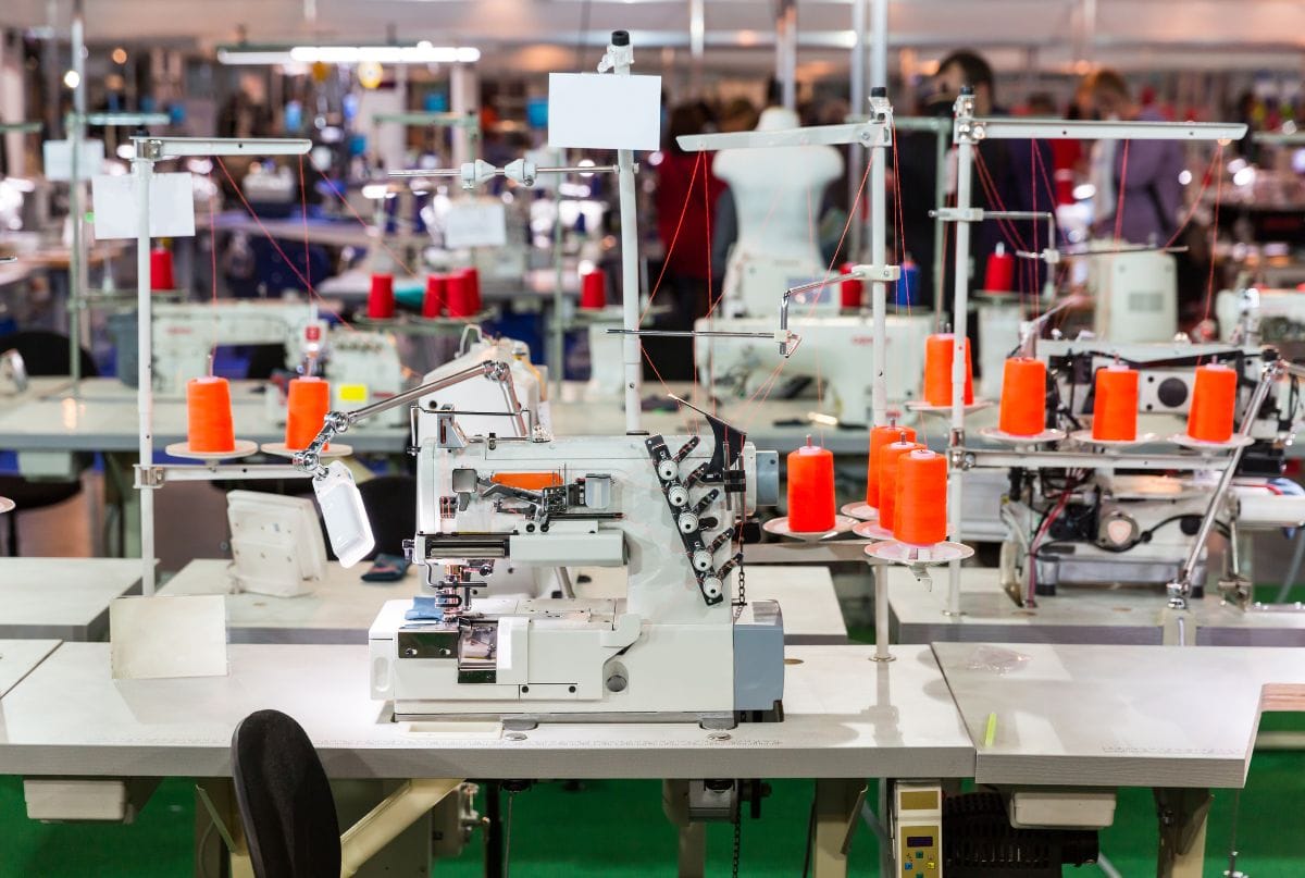 Factory sewing machines with orange thread spools, illustrating clothing manufacturing in a production facility.