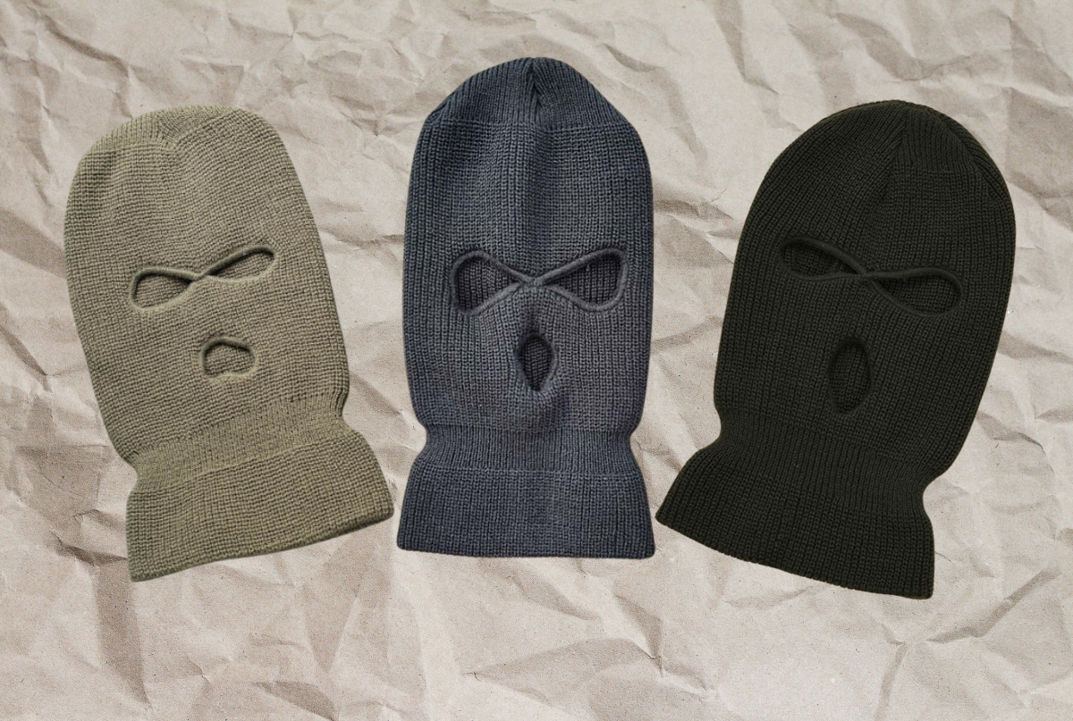 Three knitted balaclavas in different colors (beige, gray, black) arranged side by side on a crumpled paper background. Each balaclava features cut-out eye and mouth openings.