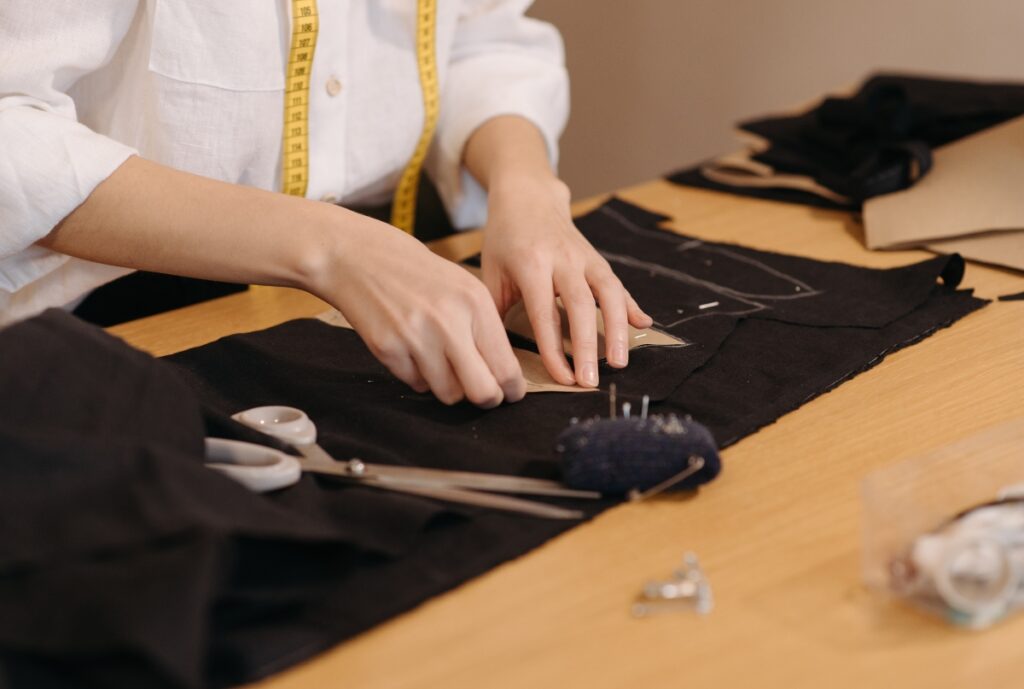 A tailor is working on customizing a black fabric on a wooden table, using scissors and other sewing tools, with a measuring tape draped around their neck.