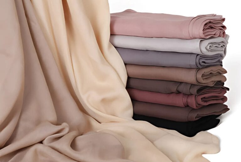 Stack of folded plain viscose hijabs in beige, pink, grey and black beside draped beige fabric