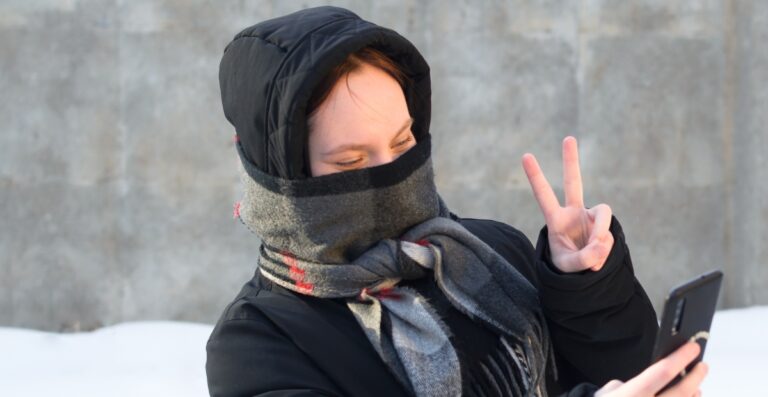 Eine Person, die in Winterkleidung gehüllt ist, einen dicken Schal trägt, der wie eine Sturmhaube um die untere Gesichtshälfte gewickelt ist, eine Kapuze und einen Mantel trägt; sie befindet sich im Freien im Schnee, hält ein Smartphone in der Hand und macht mit der anderen Hand ein Peace-Zeichen.